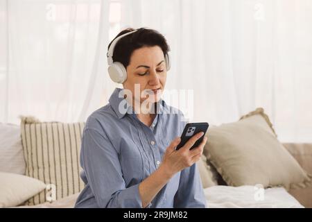 Pensive Frau mittleren Alters mit kurzen dunklen Haaren im blauen Hemd hält ihr Smartphone in der Hand und schaut auf den Bildschirm, während sie Musik mit kabellosen Kopfhörern hört Stockfoto