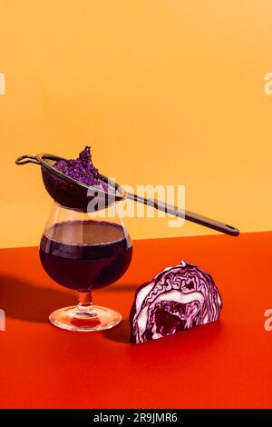 Gesundes veganes Antioxidationsgetränk hergestellt mit extrahiertem Rotkohlsaft in Glas mit Sieb auf rotem und gelbem Hintergrund Stockfoto