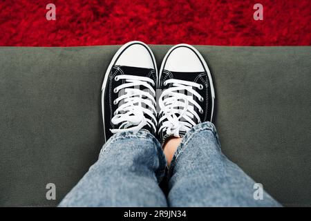 Die Beine des Mädchens sind mit klassischen Turnschuhen ausgestattet. Ein Turnschuh am Bein eines Mädchens. Teenager Füße in lässigen neuen Sneakers auf dem Sofa Nahaufnahme Bild. Vintage-Style Stockfoto