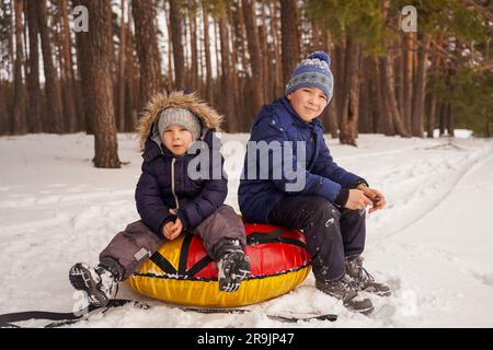 Kinder sitzen auf Tubing im verschneiten Wald und machen eine Pause vom Winterspaß. Glückliche Kinder im Winter. Stockfoto