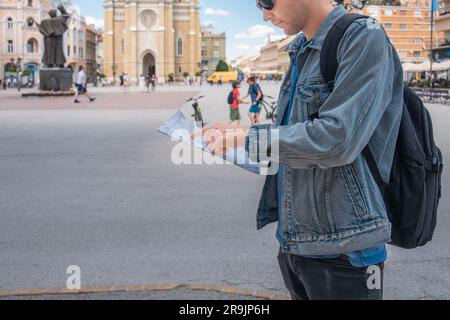 Ein Tourist auf dem Platz in der Stadt, der auf einer Karte aus Papier steht, erforscht die neue Stadt. Hübscher männlicher Reisender Rucksacktourist mit Sonnenbrille Stockfoto