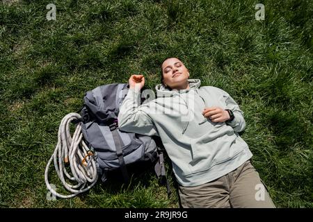 Draufsicht einer jungen, kurzhaarigen Wanderer in lässiger Kleidung, die mit geschlossenen Augen neben dem Rucksack liegt, mit Reiseausrüstung auf einem grasbedeckten Rasen, allein wandern jo Stockfoto