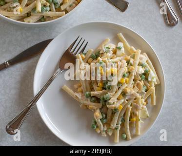 Teller mit klassischem Nudelsalat. Mit Mayonnaise, gekochten Eiern, grünen Erbsen, Mais und Wurst auf einem Tisch Stockfoto