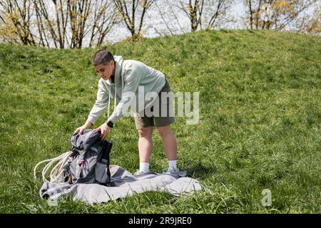 Junge, kurzhaarige Reisende Frau mit Fitness-Tracker legt Rucksack mit Reiseausrüstung auf die Decke, während sie auf einem grasbedeckten Rasen mit Hügel bei b steht Stockfoto