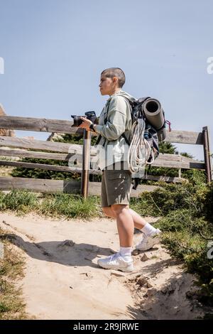 Seitenansicht einer jungen, kurzhaarigen Wanderin mit Rucksack und Reiseausrüstung, die eine Digitalkamera hält und auf einem Hügel mit dem Himmel im Hintergrund steht, Hi Stockfoto