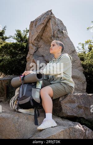 Junge Frau mit kurzen Haaren in lässiger Kleidung, die neben dem Rucksack sitzt, mit Karte auf Steinen auf einem Hügel mit Natur im Hintergrund, ruhige Wanderer, die innere sportarten finden Stockfoto