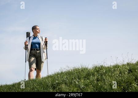 Junge, kurzhaarige Wanderer in lässiger Kleidung mit Rucksack, der Wanderstöcke hält und im Hintergrund auf einem grünen Hügel und am Himmel steht, Entdecker woma Stockfoto