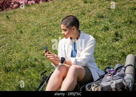 Junger, kurzhaariger und tätowierter Wanderer in legerer Kleidung mit Smartphone, während er in der Nähe von Wanderstöcken und Rucksack auf einem grünen Hügel sitzt und sich entspannt Stockfoto