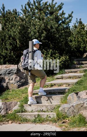 Junge Wanderer in Baseballmütze und lässiger Kleidung mit Rucksack und Reiseausrüstung, die auf Treppen gehen, mit Natur und Himmel im Hintergrund, neugierig Stockfoto