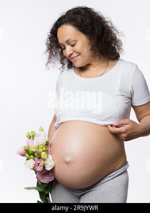 Wunderschöne, multiethnische, gravide Frau, die einen Blumenstrauß in der Hand hält, lächelt und ihren schwangeren Bauch in der späten Schwangerschaft ansieht Stockfoto