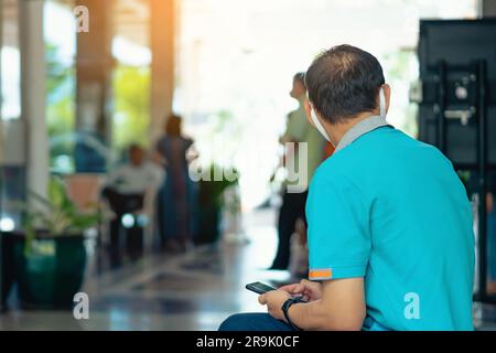 Rückansicht männlicher Reisender tragen Gesichtsmaske sitzen Sie, um Ihr Smartphone zu benutzen, während Sie auf den Hoteleincheckvorgang in der Hotellobby warten. Geschäftsmann, der in der Hotellobby wartete Stockfoto