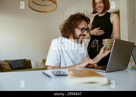 Ein junger lächelnder Mann, der von zu Hause aus an einem Laptop arbeitet, während ihm die Freundin Kaffee bringt Stockfoto