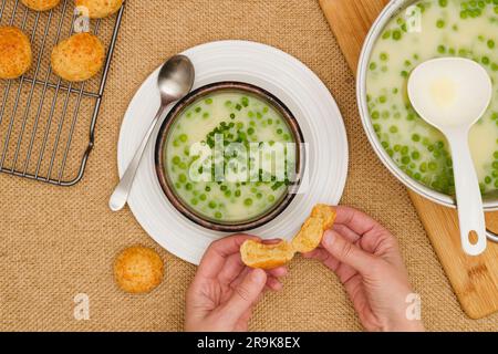 Cremige pürierte Selleriesuppe mit grüner Erbse. Frisch zubereitete Gemüsesuppe in einer Schüssel, serviert mit frisch gebackenem Käsebrot, Nahaufnahme, flach liegend, Frauenhände Stockfoto