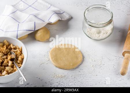 Rundes, flaches Stück gerollten Teigs, Apfelfüllung mit Zimt, Mehl und Rollnadel auf einem hellgrauen Tisch. Köstliche Mini-Pasteten mit Apfel Stockfoto