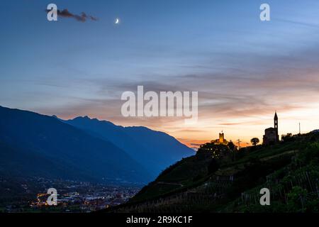 Mondschein bei Sonnenuntergang über den Silhouetten von Castel Grumello und der alten Kirche auf dem Hügel, Valtellina, Sondrio, Lombardei, Italien Stockfoto