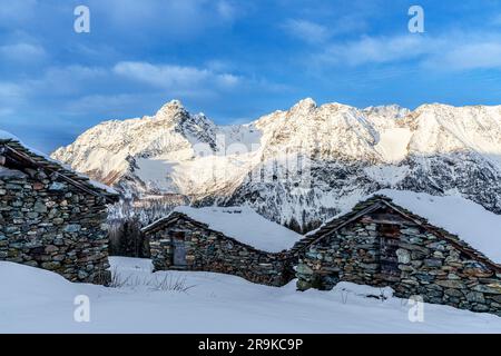 Schneebedeckte Berghütten mit schneebedeckten Gipfeln im Winter, Entova Alp, Valmalenco, Valtellina, Lombardei, Italien Stockfoto