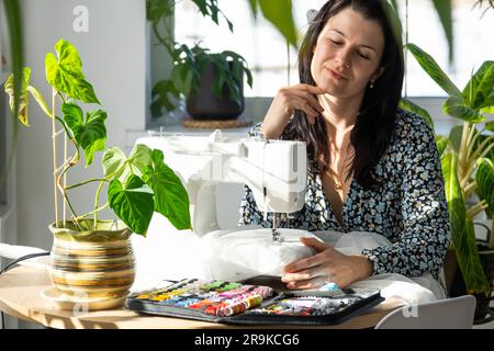 Eine Frau, die denkt, träumt und näht eine elektrische Nähmaschine in einem weißen, modernen Innenraum eines Hauses mit großen Fenstern und Zimmerpflanzen. Komfort Stockfoto