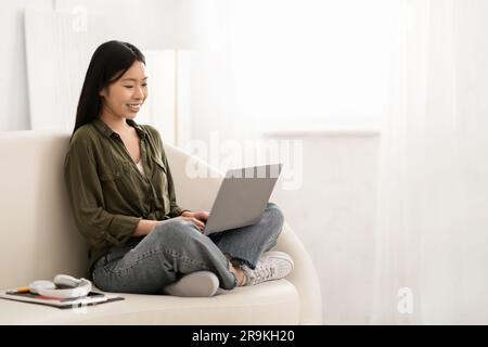 Lächelnde chinesin, die auf der Couch mit Computer sitzt Stockfoto