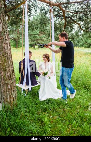 Ein professioneller Hochzeitsvideograf dreht einen Film mit den Frischvermählten auf einer Seilschaukel in einem Pinienwald Stockfoto