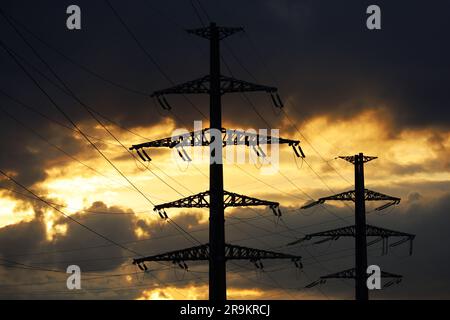 Silhouetten eines Hochspannungsturms mit elektrischen Drähten auf dem Hintergrund des Sonnenuntergangs und dunkler Wolken. Stromübertragungsleitungen, Stromversorgung Stockfoto