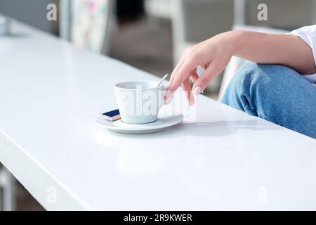 Die Hand eines Mädchens mit Maniküre nimmt einen Becher mit einem Drink. Ein Mädchen in einem Café trinkt eine Tasse Kaffee in der Hand in einem Sommerrestaurant. Weiße Tasse C. Stockfoto