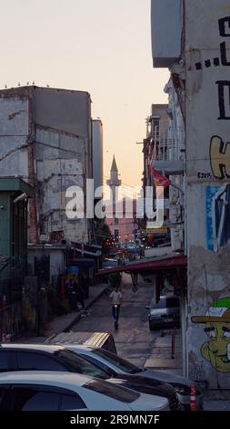 Der Mann fährt mit einem Roller mitten in einer schmalen, heruntergekommenen Straße bei Sonnenuntergang mit einem Minarett hinter dem man sehen kann. Istanbul, Türkei Stockfoto
