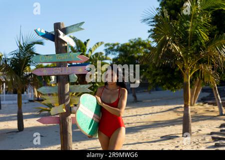 Glückliche birassische Frau in rotem Badeanzug mit Surfbrett am sonnigen Strand Stockfoto