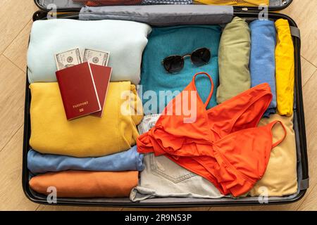 Gefaltete Damenbekleidung, Pässe mit Dollars, Badeanzug und Sonnenbrille in einem Koffer auf Holzhintergrund, von oben nach unten. Verpackung für Fahrt bei Stockfoto