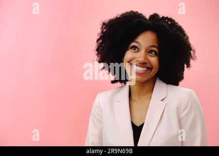 Porträt einer jungen schwarzen Frau mit pinkfarbener Jacke, die lächelt und aufblickt, vor pinkfarbenem Studiohintergrund Stockfoto