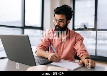 Porträt eines bärtigen indischen Geschäftsmanns mit Brille, der mit einem Laptop Projektergebnisse analysiert und Notizen an einem Tisch an einem Fenster schreibt. Stockfoto