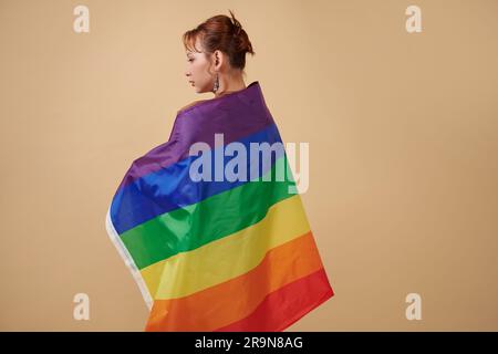 Eine echte Transgender-Frau, eingewickelt in eine Regenbogenfahne, die vor beigem Hintergrund steht Stockfoto