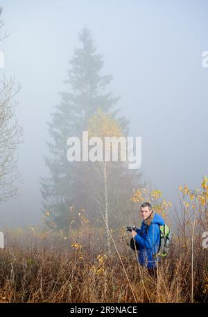 Männlicher Reisender mit professioneller Kamera, der auf einem grasbedeckten Feld mit nebligen Bäumen und Nebel im Hintergrund steht Stockfoto