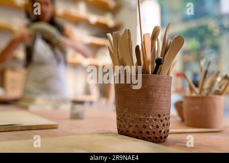 Becher mit schmutzigen Kunstwerken und handwerklichen Formwerkzeugen auf Holztisch in Töpferwerkstatt, für die Arbeit mit Ton und Keramik Stockfoto