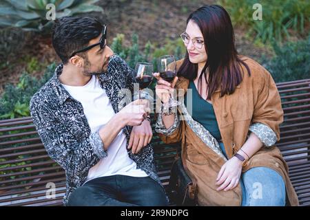 Ein junges Paar, ungezwungen gekleidet, trinkt mit einem Glas Rotwein auf einer Parkbank, und tauscht Lächeln und Blicke aus. Stockfoto