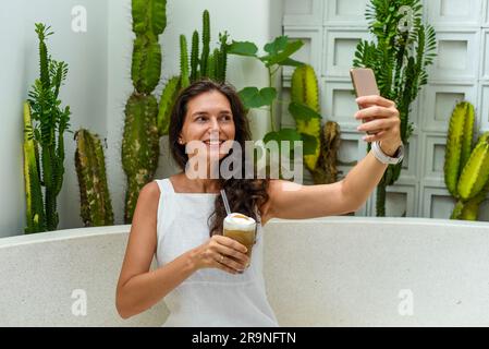 Porträt einer 39-jährigen russin, die sich nicht schminkt, mit einem Glas Eiskaffee in weißem Kleid sitzt und ein Selfie macht Stockfoto