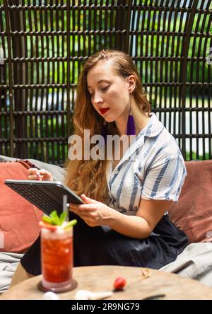 Eine wunderschöne blonde Künstlerin und Illustratorin, die über ein Konzept ihrer Zeichnung nachdenkt und mit einem elektronischen Tablet und Eingabestift am Tisch sitzt Stockfoto