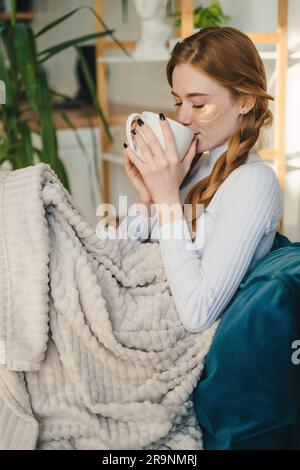 Seitenansicht einer jungen Frau in legerer Kleidung, die auf dem Sofa sitzt und heißes Getränk trinkt, während sie sich in einem gemütlichen Zimmer zu Hause ausruht Stockfoto