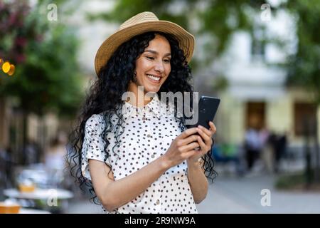 Eine wunderschöne junge Frau läuft mit einem Hut durch die Abendstadt, eine lächelnde lateinamerikanische Frau hält ein Smartphone in der Hand. Ein Tourist mit lockigem Haar tippt eine Nachricht und durchsucht Online-Seiten am Telefon. Stockfoto