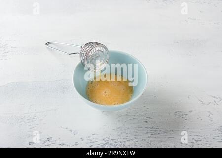 Eine blaue Schüssel mit einem geschlagenen Ei und einem Schneebesen auf einem hellblauen Tisch. Stufe der Selbstbacke. Stockfoto