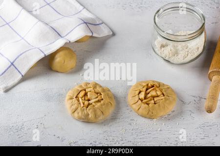 Rohe halbfertige Mini-Pasteten oder Vatrushka mit Äpfeln und Zimt, ein Glas Mehl und eine Walznadel auf einem hellgrauen Tisch, Draufsicht. Die Küche ist lecker Stockfoto