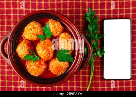 Hähnchenfleischbällchen in Tomatensoße und Smartphone mit weißem, leerem Bildschirm Stockfoto