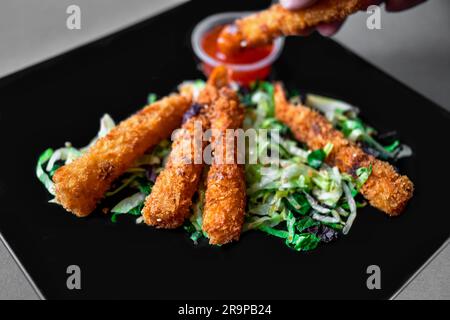 Gebratene panierte Garnelen auf Gemüse und Kräuterblätter auf schwarzem Teller, Nahaufnahme. Die Garnelen werden von Hand in scharfe Sauce getaucht. Stockfoto