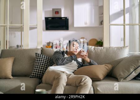 Junger asiatischer Erwachsener, der auf der Couch liegt und zu Hause auf das Handy schaut, Konzept für Smartphone- oder Social-Media-sucht Stockfoto