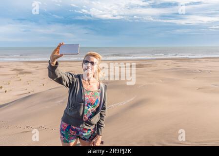 Reife Frau, die am Strand lächelt, während sie mit dem Handy Selfies macht. Sommer-, Reise- und Technologiekonzept. Stockfoto