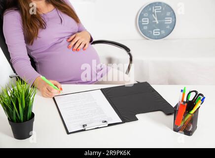 Ein schwangeres Mädchen im Büro unterschreibt Dokumente. Der Begriff der Schwangerschaft und des Elternurlaubs. Rechte und Pflichten schwangerer Frauen, Stockfoto