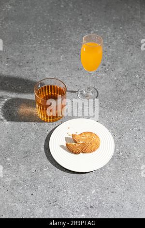 Ein weißer Teller mit frisch gebackenen Plätzchen auf einer grauen Betonoberfläche, mit zwei Gläsern bunter, erfrischender Getränke daneben Stockfoto