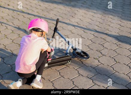 Ein 7-jähriges Mädchen mit rosa Helm und Schutzausrüstung fiel vom Roller. Das Konzept des sicheren Fahrens mit einem Roller und einem Fahrrad ohne Verletzungen. Stockfoto