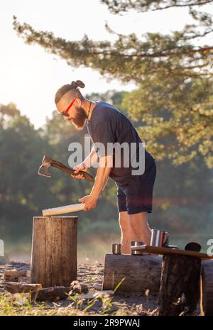 Ein eleganter, brutaler Mann mit Bart und schwarzer Brille hackt Holz auf einem Campingplatz, um ein Feuer anzünden. Outdoor-Aktivitäten im Sommer. Stockfoto