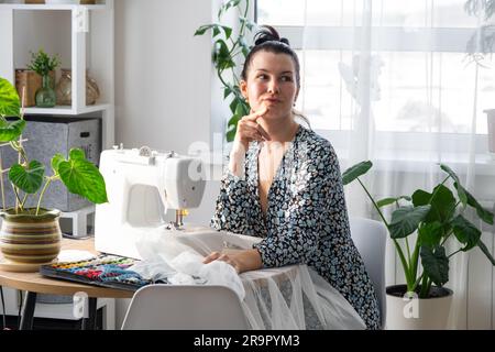 Eine Frau, die denkt, träumt und näht eine elektrische Nähmaschine in einem weißen, modernen Innenraum eines Hauses mit großen Fenstern und Zimmerpflanzen. Komfort Stockfoto