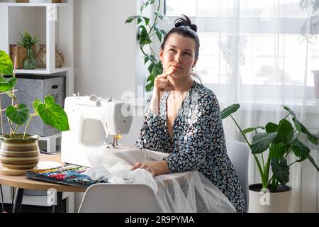 Eine Frau, die denkt, träumt und näht eine elektrische Nähmaschine in einem weißen, modernen Innenraum eines Hauses mit großen Fenstern und Zimmerpflanzen. Komfort Stockfoto